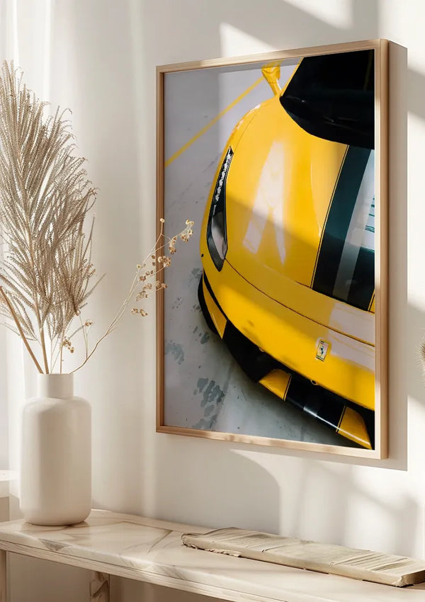 Aan een witte muur hangt een ingelijste foto van een Prachtige gele Ferrari 488 Pista met zwarte racestrepen, waardoor de voorkant van de auto goed zichtbaar is. Deze stijlvolle wanddecoratie van CollageDepot wordt gemonteerd middels een magnetisch ophangsysteem. Onder de foto staat een witte vaas met decoratieve gedroogde planten sierlijk op een lichte houten plank.,Lichtbruin