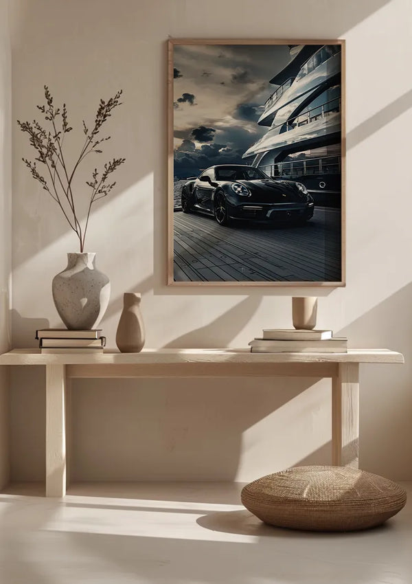 Een minimalistische kamer met een houten bank met decoratieve vazen, een boek en een laag rond kussen. Boven de bank hangt een groot ingelijst Pure Luxury Schilderij van een zwarte auto geparkeerd op een kade met een jacht op de achtergrond, tegen een dramatische lucht uit CollageDepot. De wanddecoratie wordt beveiligd door een innovatief magnetisch ophangsysteem.,Lichtbruin