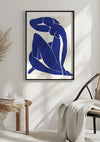 Aan een witte muur hangt een ingelijst Abstract Blauwe Vrouwelijke Silhouet Schilderij van CollageDepot met een blauw silhouet van een zittende figuur. Beneden staat een houten tafel met een glas water en gestapelde boeken naast een rieten stoel met kussens en een lichtgekleurde deken eroverheen gedrapeerd, wat een minimalistische uitstraling biedt.,Zwart