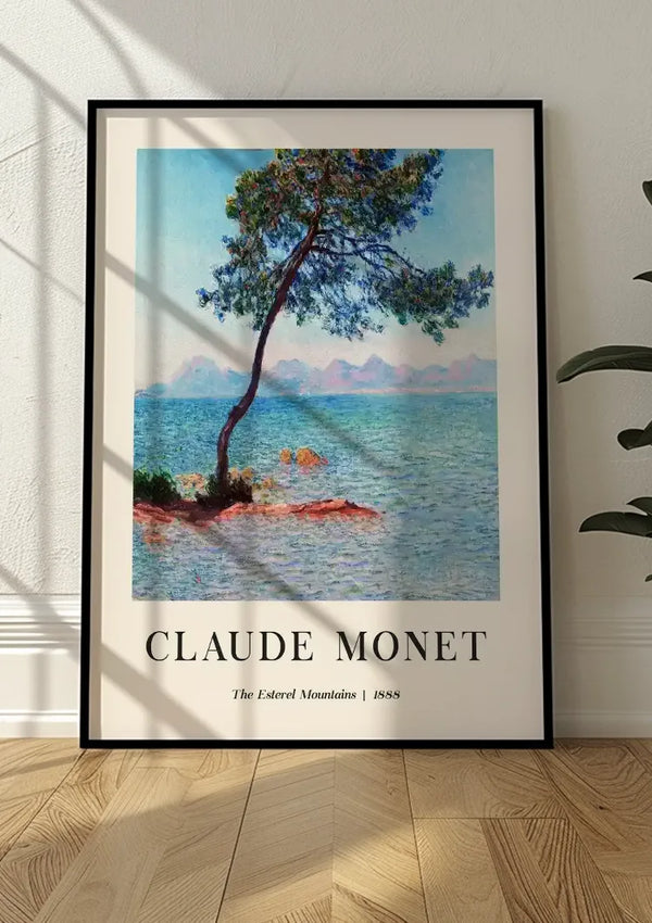 Een ingelijste kunstafdruk van Claude Monet's "The Esterel Mountains" uit 1888 rust tegen een witte muur op een houten vloer. Het schilderij, een prachtig CollageDepot Claude Monet The Esterel Mountains Schilderij, toont een boom die over water leunt met bergen op de achtergrond. Het frame werpt een schaduw op de muur en de vloer.,Zwart