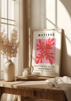 Een ingelijst CollageDepot Matisse The Cut-Outs" 1953 Schilderij met rode abstracte vormen leunt tegen een beige muur op een houten tafel en dient als opvallende wanddecoratie. Op de tafel staan ook een beige doek, een stapel boeken en gedroogde planten in een vaas , dat wordt verlicht door natuurlijk zonlicht vanuit een nabijgelegen raam met vitrages.,Lichtbruin