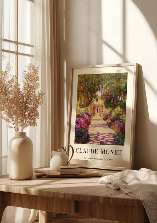 Een ingelijste poster van Claude Monet's schilderij "The Artist's Garden at Giverny" wordt als boeiende wanddecoratie op een houten tafel getoond. Op de tafel staat ook een witte vaas met gedroogde bloemen, een witte kan, een stapel boeken en een wit kleed. Het zonlicht stroomt door het raam naar binnen. Het ingelijste stuk is van CollageDepot en getiteld "Claude Monet The Artist's Garden in Giverny 1902 Schilderij.,Lichtbruin