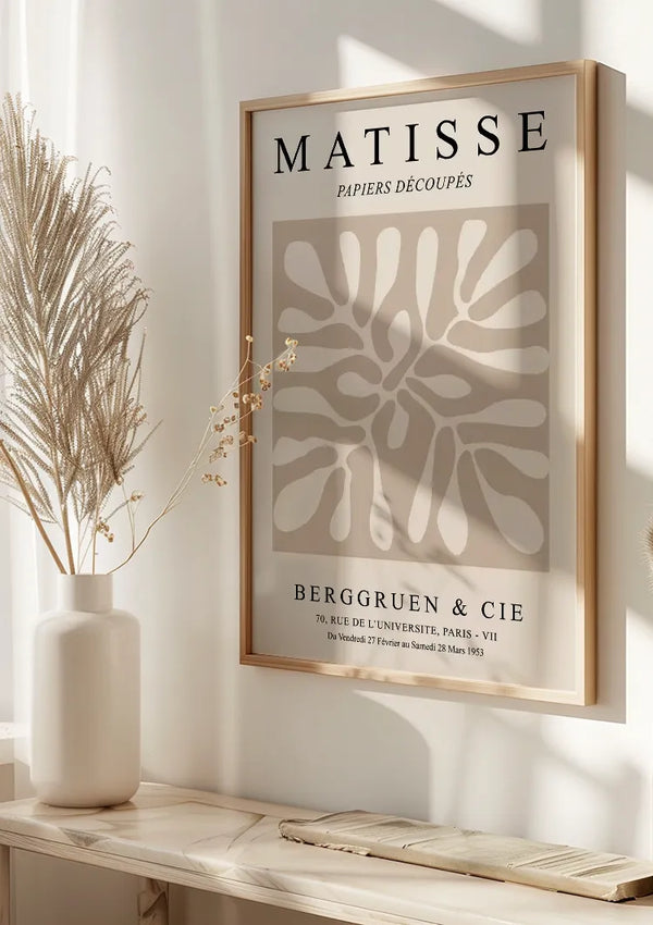 Aan de muur hangt een ingelijste poster van de *Matisse Papiers Découpés Berggruen en Cie Schilderij* uit 1953 van *CollageDepot*, beveiligd door een magnetisch ophangsysteem. De poster toont abstracte vormen in beige. Op de witte plank eronder wordt een decoratieve gedroogde plant in een vaas geplaatst, waardoor een elegante wanddecoratie ontstaat die baadt in het zonlicht.,Lichtbruin