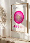 Een ingelijst Bauhaus 1919 Pink Circles-schilderij van CollageDepot hangt aan een muur, met concentrische cirkels in verschillende tinten roze en paars met bovenaan de tekst "Bauhaus 1919". De wanddecoratie is op een lichtgekleurde ondergrond geplaatst naast een minimalistische vaas met gedroogde planten, beveiligd door een magnetisch ophangsysteem.,Lichtbruin