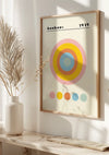Aan de muur hangt een ingelijst CollageDepot Bauhaus 1919 - Abstracte Cirkelkunst Schilderij met kleurrijke concentrische cirkels in roze, geel, blauw en grijs met kleinere cirkels aan de onderkant. De tekst bovenaan luidt "bauhaus 1919." Een witte vaas met gedroogde planten wordt met behulp van een magnetisch ophangsysteem op een tafel onder de wanddecoratie geplaatst.,Lichtbruin