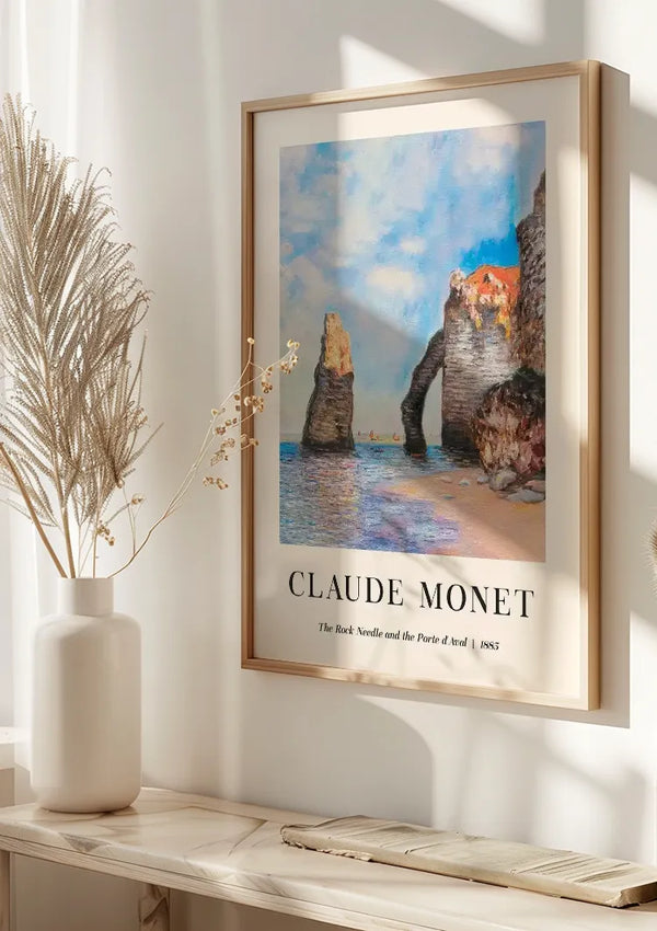 Een ingelijste Claude Monet The Rock Needle And The Porte d'Aval Schilderij poster van CollageDepot wordt op een muur getoond met behulp van een magnetisch ophangsysteem. Het kunstwerk afgebeeld op de poster is getiteld "The Rock Needle and the Porte d'Aval", gedateerd 1885. Een witte vaas met gedroogd gras wordt op een witte tafel onder de wanddecoratie geplaatst.,Lichtbruin