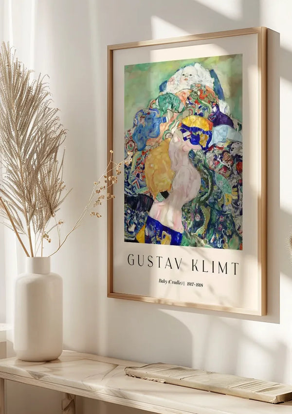 Een ingelijste poster van CollageDepot's "Gustav Klimt Baby (Cradle) Schilderij" wordt aan een helder verlichte muur gehangen. Links staat een vaas met gedroogd pampagras op een witte, minimalistische plank. Zonlicht werpt schaduwen, waardoor een elegante wanddecoratie ontstaat.,Lichtbruin