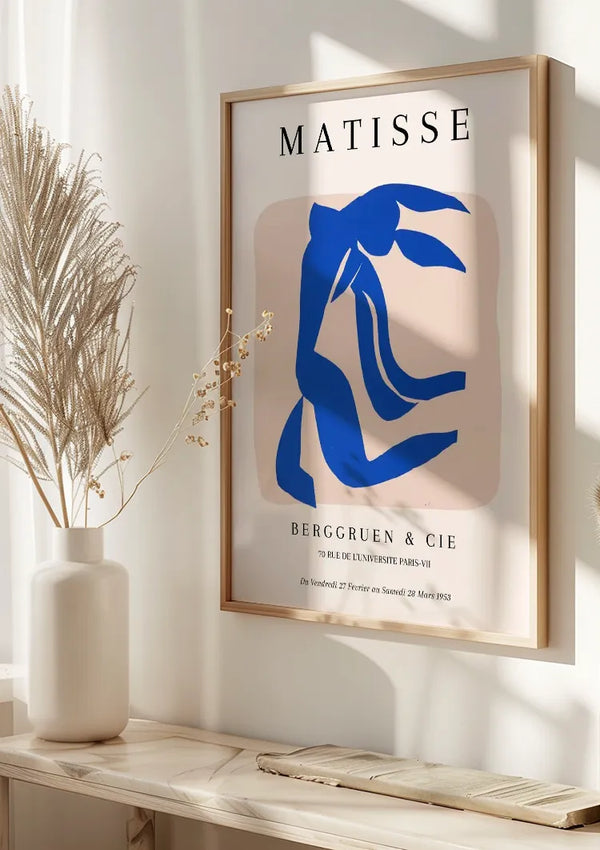 Aan de muur hangt een ingelijst Matisse Blauw Figuur Schilderij van CollageDepot, met een abstracte blauwe figuur op een lichtbruine achtergrond. De tekst eronder luidt "BERGGRUEN & CIE" met aanvullende details in kleiner lettertype. Vlakbij staat een keramieken vaas met gedroogd gras op een plank, wat de wanddecoratie prachtig accentueert.,Lichtbruin