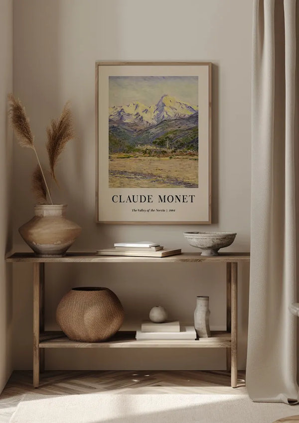 Een ingelijste print van CollageDepot’s Claude Monet The Valley Of The Nervia Schilderij dient als een prachtige wanddecoratie op een neutraal gekleurde muur boven een houten consoletafel. De tafel is versierd met vazen, een rieten mand, een kom en enkele boeken. Gedroogd riet wordt in een vaas gerangschikt.,Lichtbruin