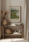 In een gezellige hoek staat een houten consoletafel met decoratieve items. Op de onderste plank staan een geweven mand en aardewerk, terwijl er bovenop een vaas met gedroogd riet en een kom staan. Hierboven hangt als wanddecoratie een ingelijst CollageDepot Claude Monet View Of Vétheuil Schilderij aan de muur.,Lichtbruin