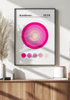 Een ingelijst Bauhaus 1919 Pink Circles-schilderij van CollageDepot met een ontwerp bestaande uit concentrische cirkels in roze tinten hangt aan een witte muur met behulp van een magnetisch ophangsysteem. Bovenaan staat "Bauhaus 1919" geschreven. Onder het schilderij bevindt zich een houten kast met diverse decoratieve spullen, waaronder vazen en boeken.,Zwart