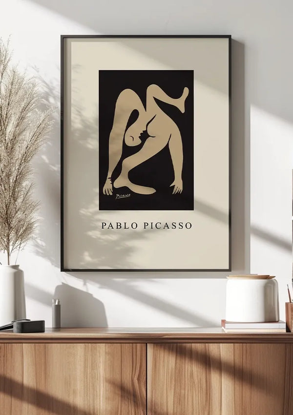 Een ingelijste poster van een kunstwerk van Pablo Picasso hangt aan een witte muur boven een bruin houten dressoir. De zwart-beige abstracte figuur wordt centraal weergegeven op het Pablo Picasso - Beige Abstract Figuur Schilderij van CollageDepot. Op het dressoir staan diverse decoratieve items, waaronder keramische vazen en een gedroogde plant, die charme toevoegen aan de wanddecoratie.,Zwart