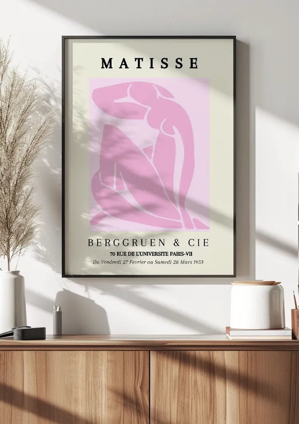 Een ingelijste kunstposter met het woord "Matisse" bovenaan, met een roze abstract figuur op een lichtbeige achtergrond. Onder de figuur luidt de tekst "Berggruen & Cie" en worden tentoonstellingsdetails in het Frans gegeven. Het frame rust op een houten plank met decoritems. Dit kunstwerk is geïdentificeerd als ccc 038 - bekend Schilderij van CollageDepot.,Zwart