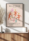 De afbeelding toont een poster "Matisse - The Dance Levendige Dansers 1953 Schilderij" van CollageDepot, ingelijst en opgehangen met behulp van een magnetisch ophangsysteem boven een houten kast. De poster toont abstracte menselijke figuren die dansen, omringd door kleurrijke organische vormen op een beige achtergrond. Op de kast staat een vaas met gedroogde planten en een boekje.,Zwart