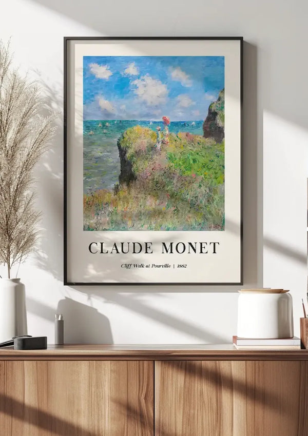 Een ingelijst CollageDepot Claude Monet - Cliff Walk at Pourville Schilderij hangt aan een witte muur boven een houten kast. Het schilderij, voorstellende groene kliffen en een blauwe lucht, is beveiligd met een magnetisch ophangsysteem. In de kast staan decoratieve spullen, waaronder een vaas met gedroogd gras.,Zwart