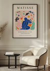 Een ingelijste Matisse Tentoonstellingposter 1953 Schilderij van CollageDepot hangt aan een beige muur en dient als elegante wanddecoratie. Het kleurrijke kunstwerk toont een blauwe abstracte figuur omringd door levendige vormen en planten. Onder het kunstwerk staat de tekst "Matisse, Berggruen & Cie" en tentoonstellingsdetails. Hieronder bevinden zich een fauteuil en een bijzettafel.,Zwart