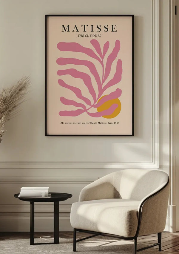 Een ingelijste poster met de titel "Matisse Roze Bladmotief Schilderij" van CollageDepot hangt aan een crèmekleurige muur boven een witte fauteuil en een klein zwart rond tafeltje. De poster, voorzien van een roze en geel abstract bladdessin, wordt bevestigd door middel van een magnetisch ophangsysteem. Een getextureerde plant siert de linkerkant van de afbeelding.,Zwart