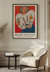 Een ingelijst CollageDepot H. Matisse La Blouse Roumaine Schilderij hangt als prachtige wanddecoratie aan een beige muur boven een moderne witte fauteuil. Naast de stoel staat een ronde zwarte bijzettafel en links is gedeeltelijk een potplant zichtbaar. Natuurlijk licht verlicht de ruimte.,Zwart