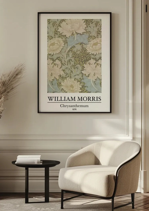 Een ingelijste CollageDepot W. Morris Chrysanthemum Schilderij met de titel "William Morris Chrysanthemum, 1876" wordt getoond op een witte muur boven een beige fauteuil. Het kunstwerk heeft een botanisch ontwerp met groene bladeren en witte bloemen. Naast de stoel staat een zwart bijzettafeltje met een stapel boeken, wat de wanddecoratie perfectioneert.,Zwart