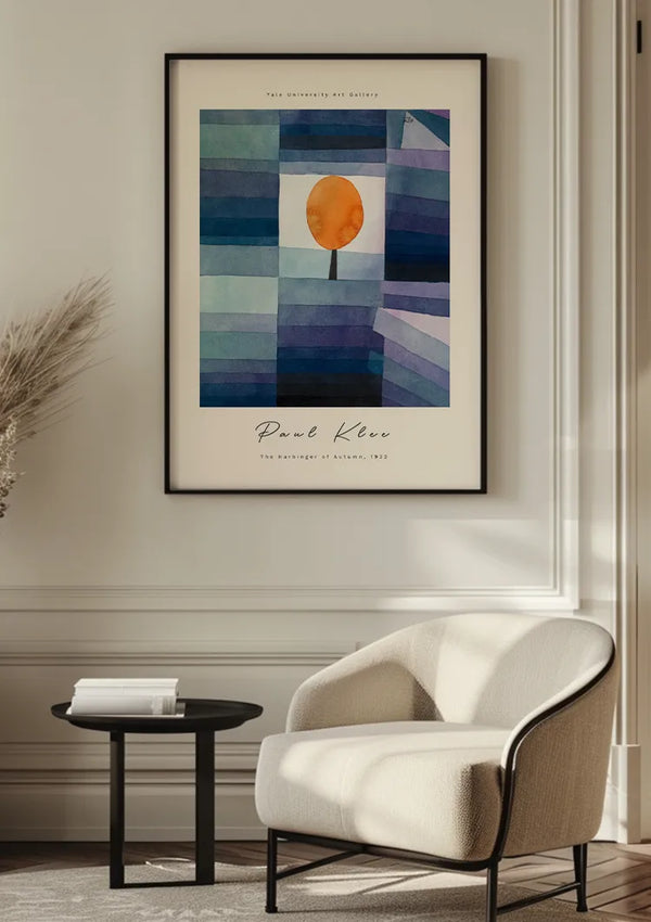 Een ingelijst CollageDepot P. Klee schilderij De voorbode van de herfst 1922 hangt aan de muur boven een witte stoel met kussens en een klein bijzettafeltje. Het kunstwerk, beveiligd door een magnetisch ophangsysteem, toont een boom met een oranje kroon tegen een geometrische achtergrond met tinten blauw en paars. De tekst van de prent luidt: "Paul Klee: The Relationships of Harmony, 1929.,Zwart