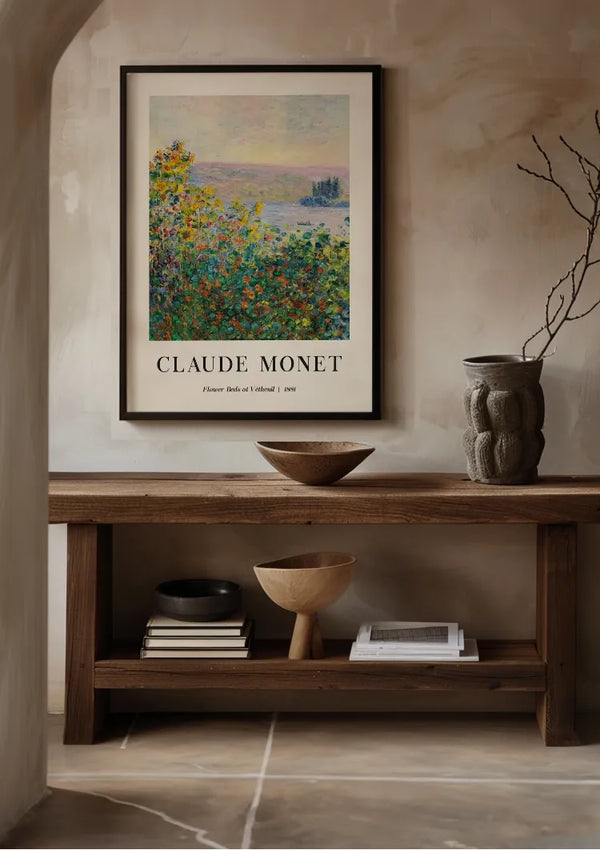 Een ingelijste Claude Monet-bloembedden bij Vétheuil Schilderij van CollageDepot in een impressionistische stijl worden boven een houten tafel getoond. Op de tafel staan schalen, een decoratieve vaas en een stapel boeken. Het schilderij toont "Bloembedden bij Vétheuil" uit 1887, in een goed verlichte, neutraal getinte kamer met behulp van een magnetisch ophangsysteem.,Zwart