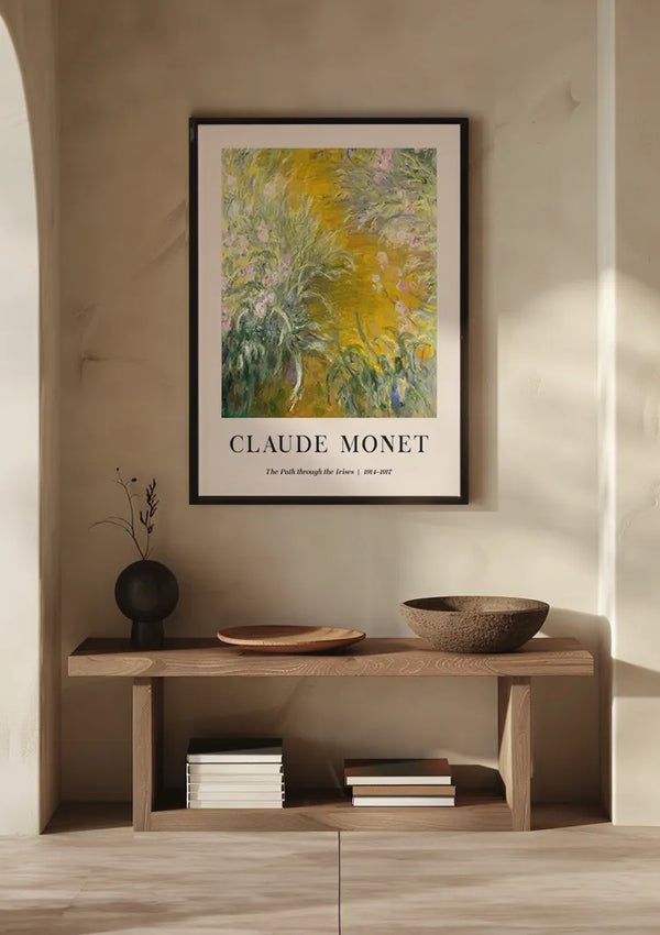Een ingelijst schilderij van Claude Monet The Path Through The Irisis van CollageDepot, getiteld "The Path through the Irises", hangt aan een beige muur boven een houten consoletafel. Op de tafel staat een vaasje met takjes, een ondiepe schaal, een houten dienblad en een stapel boeken, allemaal perfect uitgelijnd met behulp van een magnetisch ophangsysteem.,Zwart