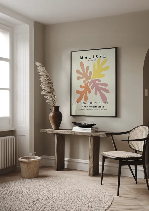Een minimalistische, neutraal getinte kamer is voorzien van een houten consoletafel met decoratieve items, waaronder een vaas met pampagras en een kom. Boven de tafel hangt een ingelijste Matisse: The Cut-Outs Berggruen & Cie 1953 Schilderij, beveiligd door een magnetisch ophangsysteem. Een geweven stoel en een rond vloerkleed maken het decor compleet.,Zwart