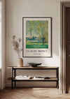 Een ingelijst Claude Monet The Willows Schilderij van CollageDepot hangt aan een witte muur boven een minimalistische houten consoletafel. Op de tafel staat een keramieken vaas met gedroogde planten, een open boek en een ondiepe schaal. De achtergrond bestaat uit een deel van een deuropening en zachte natuurlijke verlichting, wat deze charmante wanddecoratie compleet maakt.,Zwart