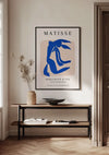 Een minimalistisch interieur met een ingelijste CollageDepot Matisse Blauw Figuur Schilderij op een beige muur boven een houten consoletafel. Op de tafel staat een arrangement met gedroogde planten, een ondiepe schaal en gestapelde boeken. De kamer, compleet met houten visgraatvloer, beschikt over elegante wanddecoratiemogelijkheden.,Zwart