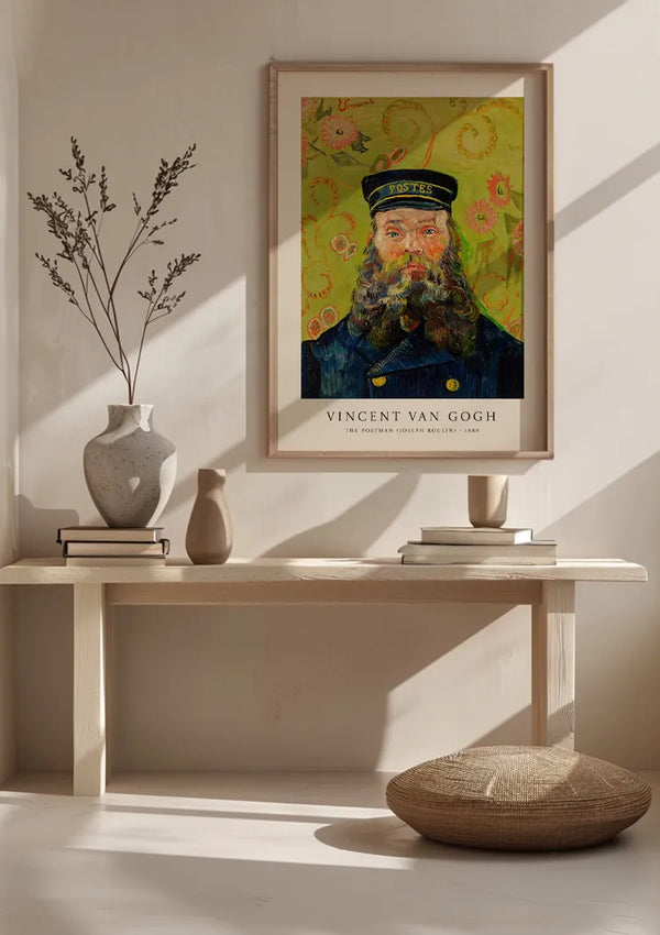 Een ingelijst CollageDepot V. Van Gogh Postbode Portret Schilderij met het schilderij "Portret van Joseph Roulin: 1889", waarop hij wordt afgebeeld in zijn postbode-uniform, hangt aan een lichtgekleurde muur boven een houten consoletafel versierd met verschillende keramische vazen, boeken, en een geweven kussen op de vloer.,Lichtbruin