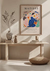 Een minimalistische kamer met een ingelijste "Matisse Tentoonstellingposter 1953 Schilderij" van CollageDepot met kleurrijke abstracte vormen en een blauwe figuur, elegant opgehangen met behulp van een magnetisch ophangsysteem boven een houten bank. Op de bank staan twee vazen, boeken en een klein decoratief object. Zonlicht werpt schaduwen op de lichtgekleurde muur.,Lichtbruin