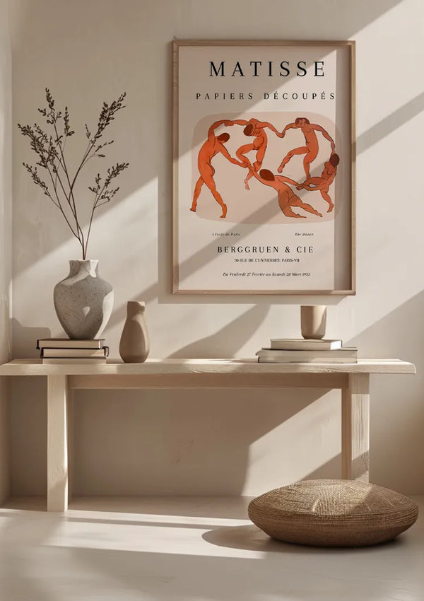 Een minimalistische kamer is voorzien van een houten tafel met een potplant, boeken en keramiek. Boven de tafel hangt een CollageDepot Matisse - The Dance Schilderij getiteld "Papiers Découpés", met een abstract ontwerp van roodoranje figuren die in een cirkel dansen. Op de vloer ligt een ronde geweven mat.,Lichtbruin