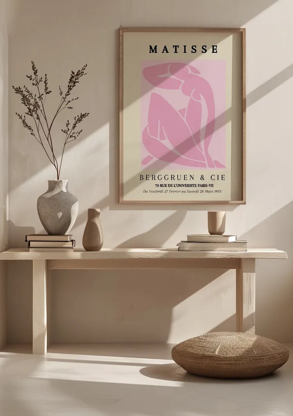 Een minimalistische houten consoletafel versierd met twee vazen met gedroogde takken, een gebeeldhouwd object en een geweven placemat staat tegen een neutrale muur. Daarboven hangt een ingelijst CollageDepot ccc 038 - bekend Schilderij met abstracte roze vormen en tekst over een kunsttentoonstelling.,Lichtbruin
