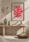 Een minimalistische muurkunstprint met een H. Matisse - Abstracte Bladvorm Schilderij met opvallende rode abstracte vormen op een roze achtergrond wordt getoond op een lichte houten plank. De wanddecoratiescène bestaat uit een witte vaas met een abstract bladvormig takje bladeren, een rond geweven kussen en zachte verlichting. Dit prachtige stuk is van CollageDepot.,Lichtbruin