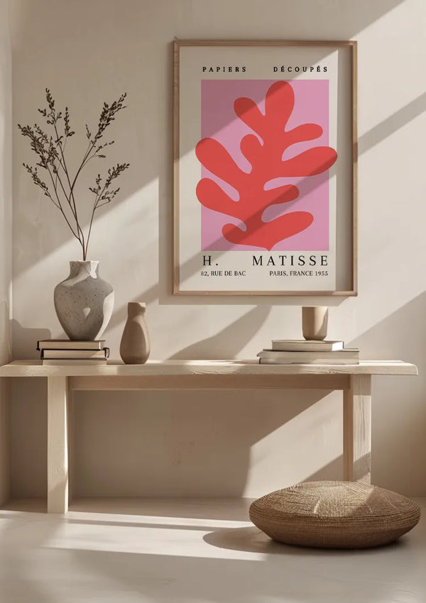 Een minimalistische muurkunstprint met een H. Matisse - Abstracte Bladvorm Schilderij met opvallende rode abstracte vormen op een roze achtergrond wordt getoond op een lichte houten plank. De wanddecoratiescène bestaat uit een witte vaas met een abstract bladvormig takje bladeren, een rond geweven kussen en zachte verlichting. Dit prachtige stuk is van CollageDepot.,Lichtbruin