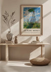 Een minimalistische decoropstelling met een ingelijste vrouw van Claude Monet met een parasol - Madame Monet And Her Son Schilderij van CollageDepot. De wanddecoratie wordt opgehangen met behulp van een magnetisch ophangsysteem boven een lichthouten bank versierd met vazen en een boek. Op de grond ligt een rond kussen en in een van de vazen staat een plantentak hoog.,Lichtbruin