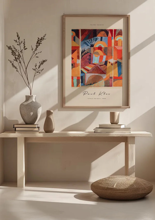 Een minimalistische kamer met een bank tegen de muur met een ingelijst abstract schilderij van Paul Klee, "Temple Gardens 1920", bewaard door een CollageDepot P. Klee Temple Gardens 1920 Schilderij magnetisch ophangsysteem. Op het bankje staan diverse vazen en gestapelde boeken. Op de grond ligt een rond kussen. De kamer wordt verlicht door natuurlijk licht, waardoor schaduwen op de muur vallen.,Lichtbruin