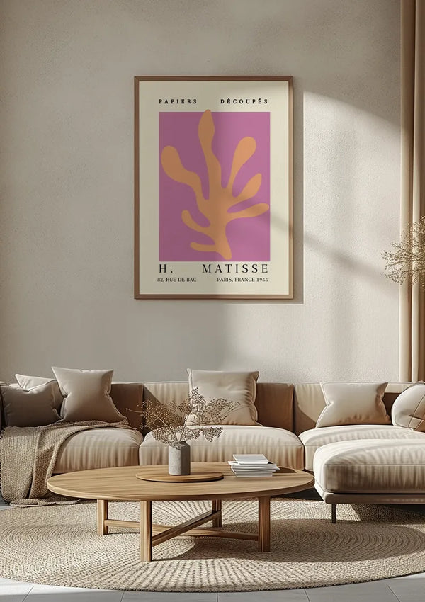Een ingelijst CollageDepot H. Matisse schilderij uit 1955 getiteld "Papiers Découpés" hangt aan een beige muur boven een eigentijdse beige bank met witte en beige kussens. Deze elegante wanddecoratie complementeert de ronde houten salontafel met een plant en boeken en maakt de serene sfeer perfect af.,Lichtbruin