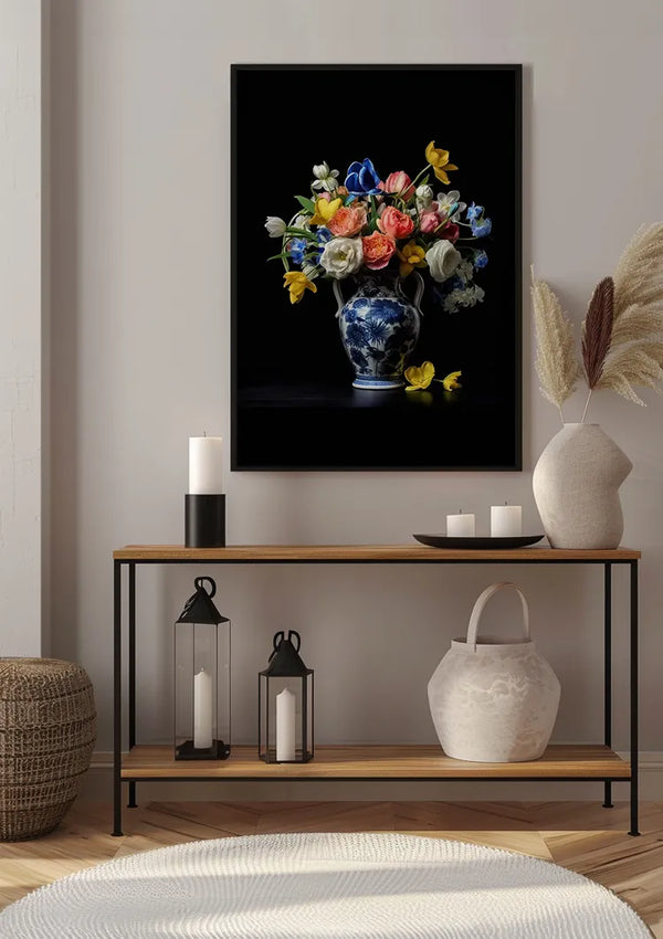 Een ingelijste CollageDepot Delfts Blauw Bloemen Vaas Schilderij van kleurrijke bloemen hangt aan een muur boven een consoletafel. Op de tafel staan twee witte stompkaarsen, lantaarns, een witte mand en een decoratief rietarrangement. Op de houten vloer worden een geweven mand en een wit rond vloerkleed geplaatst.,Zwart