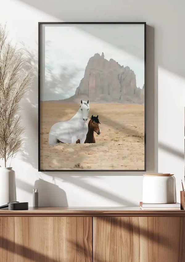 Een ingelijste foto, de perfecte wanddecoratie, hangt aan een witte muur en toont Paarden in Landschap Schilderij van CollageDepot staande in een woestijnlandschap met een rotsachtige bergformatie op de achtergrond. De foto bevindt zich boven een houten consoletafel versierd met decoratieve items.,Zwart