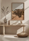 Een minimalistische kamer met een houten consoletafel met drie vazen, een ingelijste foto van olifanten die over een onverharde weg lopen, en een geweven kussen op de vloer. Een Olifanten in Beweging Schilderij van CollageDepot hangt met behulp van een magnetisch ophangsysteem, waarbij zonlicht schaduwen werpt op de lichtgekleurde muren en tafel.,Lichtbruin