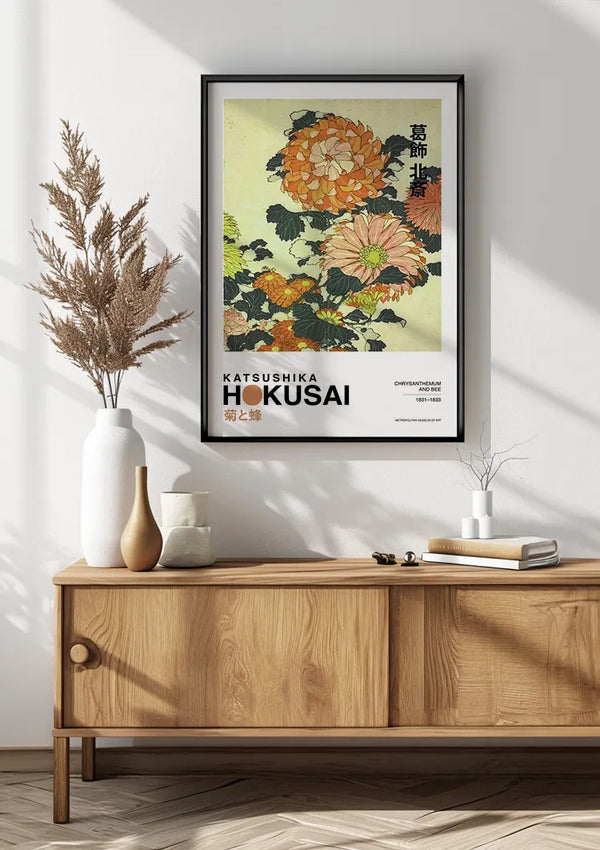 Een ingelijst CollageDepot K. Hokusai Chrysanthenum en bijenschilderij op een lichtgele achtergrond hangt aan een witte muur boven een houten dressoir. Het dressoir heeft een witte vaas met gedroogde bloemen, een kleiner keramiekstuk en twee op elkaar gestapelde dichte boeken. Deze prachtige wanddecoratie maakt gebruik van een magnetisch ophangsysteem voor eenvoudige weergave.,Zwart