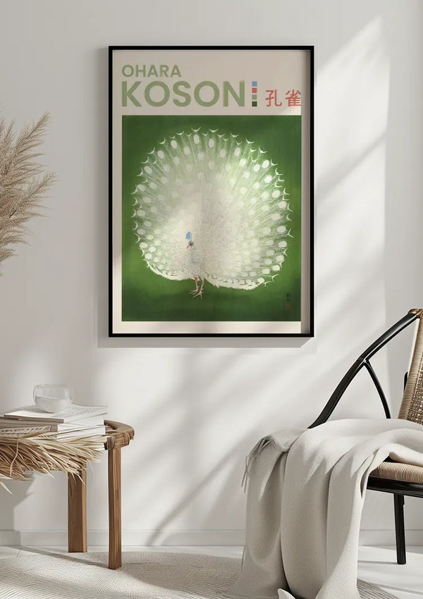 Een ingelijst O. Koson Peacock Print Schilderij van CollageDepot tegen een groene achtergrond, met de tekst "OHARA KOSON", weergegeven op een muur. Een lichtgekleurde deken wordt over een geweven stoel in de buurt gedrapeerd, terwijl op een bijzettafel een stapel boeken en een dienblad met een klein voorwerp staan.,Zwart