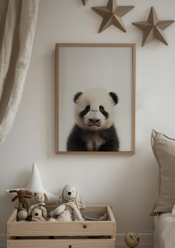 Een gezellige kamer is voorzien van een ingelijst Panda Welpje Schilderij van CollageDepot dat aan een witte muur hangt, versterkt door een stervormige wanddecoratie erboven. Onder het kunstwerk staat een houten kist gevuld met knuffels, waaronder een beer, konijn en hond. Rechts ligt een beige kussen.,Lichtbruin