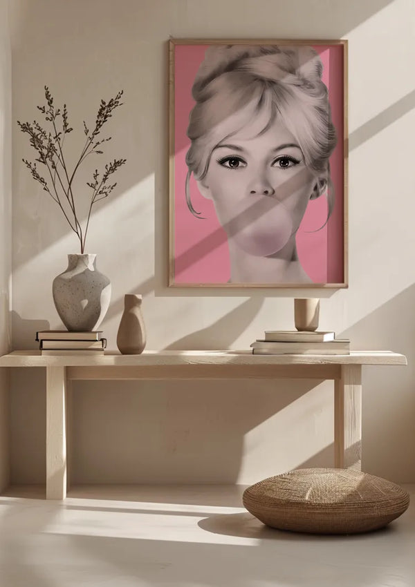 Een minimalistische kamer met een houten bank met een ingelijst Bubble Gum Bliss Popart Schilderij van CollageDepot met een zwart-witfoto van een vrouw die een kauwgombel blaast tegen een roze achtergrond. Het decor bestaat uit wanddecoratie, twee vazen en gedroogde takken. Een rond kussen ligt op de vloer terwijl het zonlicht naar binnen stroomt.