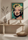 Een ingelijst Marilyn Monroe Karakteristiek Popart Portret Schilderij van CollageDepot hangt als opvallende wanddecoratie boven een houten consoletafel. Op de tafel staan twee siervazen en een stapel boeken, terwijl daarvoor een rond geweven zitkussen op de grond ligt.