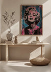 Een minimalistische kamer is voorzien van een houten consoletafel met decoratieve items, waaronder een vaas met takken, een witte vaas en gestapelde boeken. Boven de tafel hangt een CollageDepot Marilyn Monroe Spetterende Expressie Popart Schilderij beveiligd door een innovatief magnetisch ophangsysteem. Op de grond ligt een rond kussen.