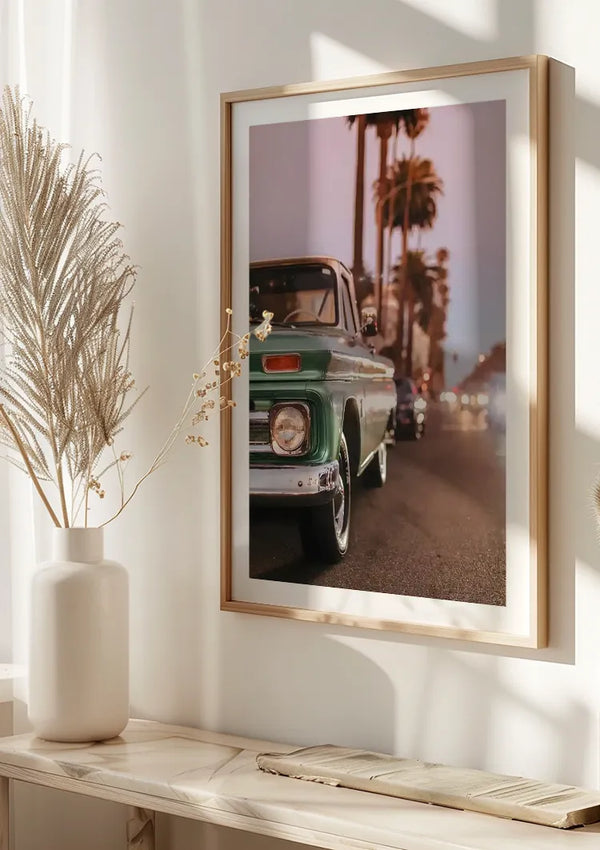 Aan een crèmekleurige muur hangt een ingelijste foto van een vintage groene vrachtwagen, geparkeerd in een straat vol palmbomen. Het Groene Oldtimer Met Palmbomen Schilderij van CollageDepot, geaccentueerd door zonlicht uit een nabijgelegen raam, heeft een witte vaas met gedroogd blad op een plank eronder.,Lichtbruin