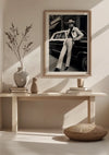 Een minimalistische kamer met lichtgekleurde houten meubels en decor. Een ingelijste zwart-witfoto van een persoon die tegen een klassieke auto leunt, dient als belangrijkste wanddecoratie boven een houten consoletafel, waarop twee vazen en een schaaltje staan. Op de grond ligt een rond kussen, aangevuld met het Lean back in style schilderij van CollageDepot.