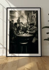 Een ingelijste zwart-witfoto leunt tegen een witte muur. De afbeelding toont drie mannen in pak, zittend rond een ronde tafel in een slecht verlichte, met rook gevulde kamer, die doet denken aan een oude bar of speakeasy. Dit "Tea time schilderij" van CollageDepot, met vage figuren op de achtergrond, is voorzien van een handig magnetisch ophangsysteem.